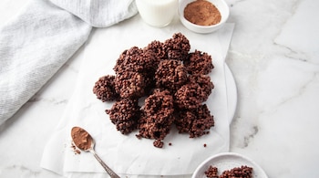 Vista aérea de bocaditos de arroz inflado con chocolate en papel de horno, un vaso de leche, cacao en polvo en un cuenco y una cuchara.