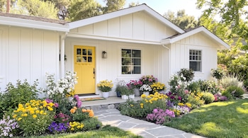 Fachada de casa blanca con puerta amarilla, camino de piedra, césped y variedad de flores de colores en el frente.