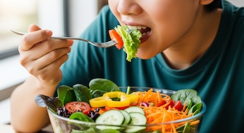 Una persona come una ensalada de vegetales frescos de un tazón de vidrio, con lechuga, tomate, pepino, zanahoria y pimiento amarillo en el tenedor.