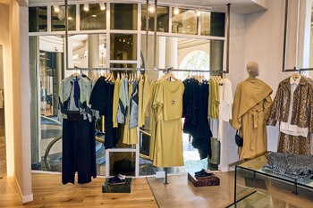 Interior de una boutique con percheros de ropa variada, maniquíes con atuendos, y un escaparate que muestra columnas y árboles del exterior