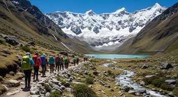 Un grupo de personas con mochilas de colores camina por un sendero rocoso, junto a un río, hacia una laguna turquesa al pie de montañas nevadas bajo cielo azul.
