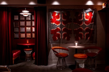 Interior de un lounge con cortinas rojas, una mesa roja y sillas naranjas. Una obra de arte abstracta roja y gris decora la pared. Iluminación tenue