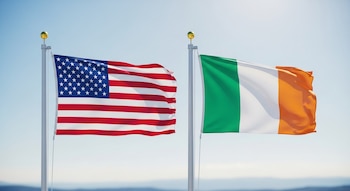 La bandera de Estados Unidos con estrellas y barras, y la bandera de Irlanda con franjas verde, blanca y naranja, ondean en mástiles bajo un cielo azul.