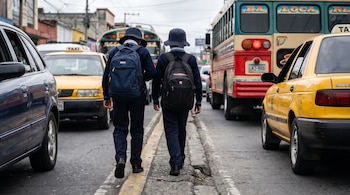 Dos niños de escuela con uniformes y mochilas caminan por una calle de Guatemala con tráfico. Hay taxis amarillos, un coche azul y un autobús.