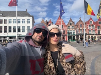 Hombre con sudadera gris y gafas de sol y mujer con abrigo de leopardo y gorro beige, posan. Fondo con edificios históricos, banderas y cielo azul
