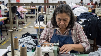 Una mujer con camisa a cuadros y delantal de mezclilla opera una máquina de coser industrial. Hay hilos y otras máquinas con personas al fondo.