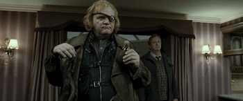Un hombre de mediana edad con cabello rubio, un ojo protésico y un frasco oscuro, vestido con un abrigo y chaleco de cuero, está en un interior con cortinas y lámparas