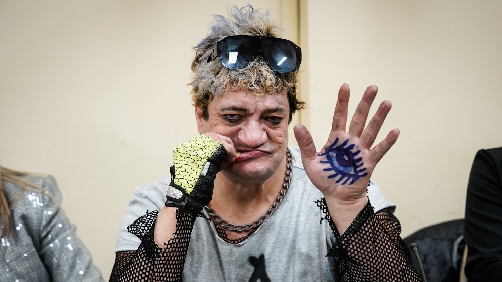 Una persona caracterizada con gafas de sol sobre la cabeza y guantes de red muestra la palma de su mano izquierda con un ojo azul dibujado.