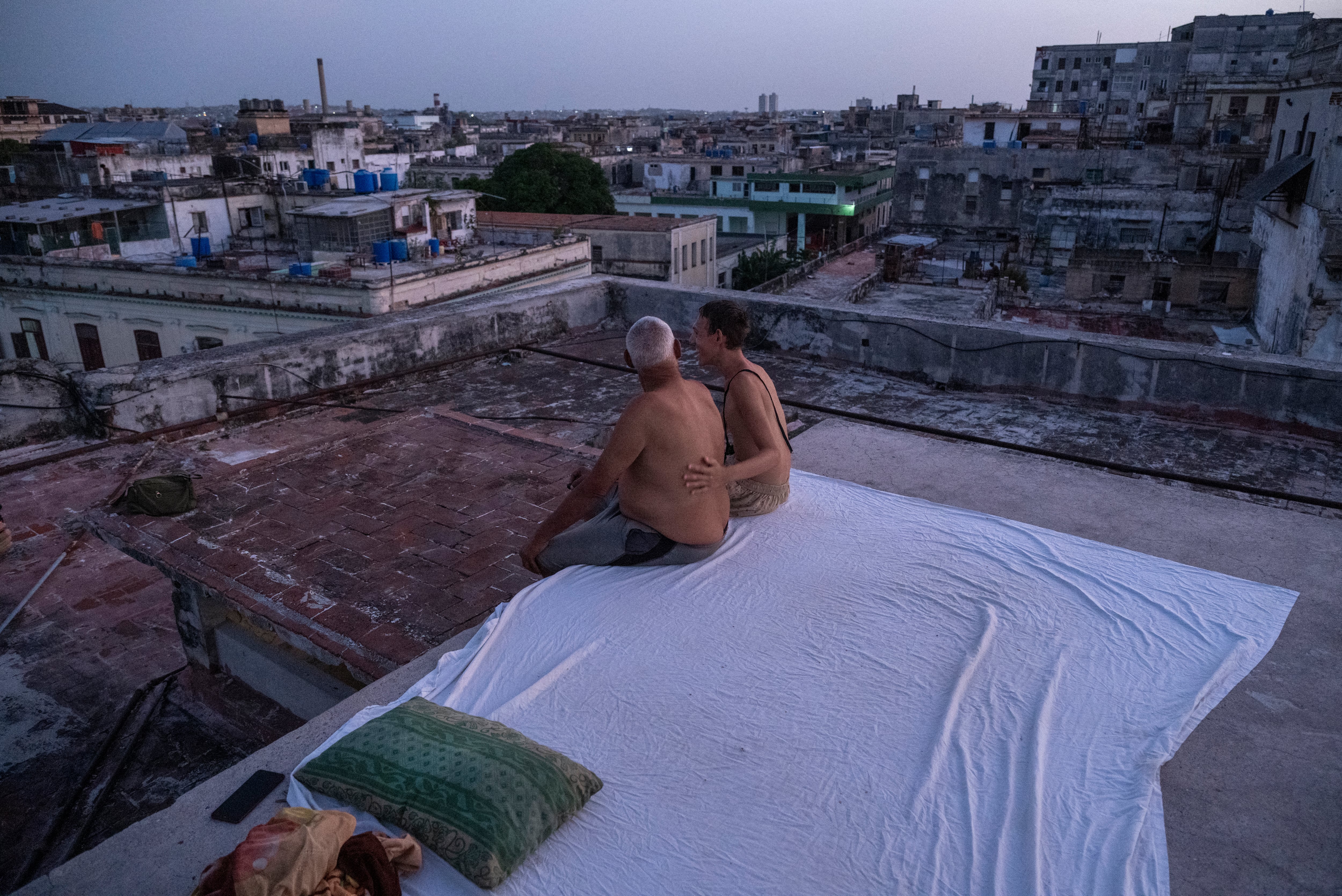 Alexis Miján y su hijo, Bayrón Miján, se sientan en una cama improvisada en la azotea de un edificio residencial al atardecer en La Habana Vieja, el 11 de agosto de 2026. Muchos cubanos duermen al aire libre durante las noches de verano para resguardarse de las altas temperaturas y los frecuentes cortes de luz. (Foto de YAMIL LAGE / AFP)