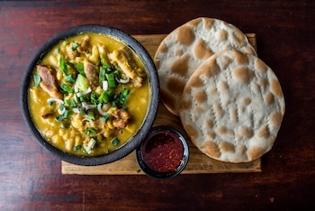 Vista aérea de un cuenco negro lleno de locro amarillo con trozos de carne y cebollino verde picado, acompañado de dos panes planos y una salsa roja