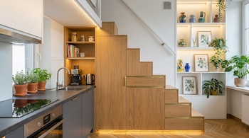Una cocina moderna con encimera oscura y fregadero de acero, adyacente a una escalera de madera clara con armarios y cajones integrados. A la derecha, estanterías blancas con decoración y plantas.