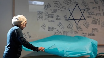 Una persona con boina beige y camisa oscura extiende una tela azul sobre una mesa, con un tablero blanco detrás que muestra una Estrella de David azul y numerosas firmas