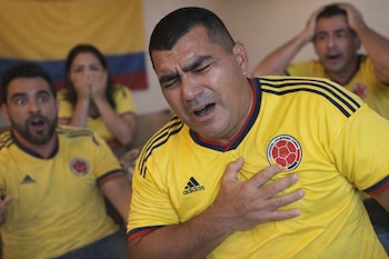 Hombre con camiseta amarilla de Colombia toca su pecho con la mano y una expresión de dolor. Al fondo, dos personas con camisetas similares muestran sorpresa.