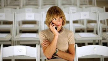 Anna Wintour, com seu característico cabelo bob, falando ao telefone com os olhos fechados, cercada por cadeiras brancas vazias