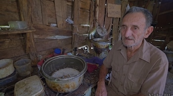 Un hombre mayor sentado en una habitación de madera junto a una olla con arroz y un cartel de cartón con el texto SIN ALIMENTOS.