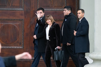 Una mujer con cabello castaño, traje de terciopelo negro y blusa blanca sonríe y saluda, saliendo por una puerta de madera oscura, acompañada por tres hombres