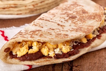 Una tortilla de harina doblada rellena de frijoles refritos, huevo revuelto y queso sobre un mantel blanco y una mesa de madera