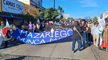 Vista frontal de una manifestación con personas llevando una gran pancarta azul que dice 'MAZARIEGO NUNCA MÁS' en una calle, con edificios al fondo