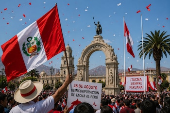 Una persona sostiene la bandera peruana y un cartel durante un acto público frente a un monumento de piedra con un arco