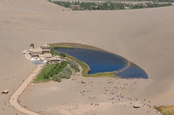 Vista aérea de un lago en forma de media luna rodeado por dunas de arena, edificios tradicionales y personas caminando sobre un camino