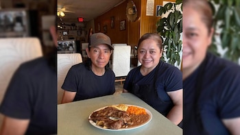 Un hombre con gorra y una mujer con delantal negro sonríen a la cámara, sentados en una mesa con un plato de carne, arroz, frijoles y huevos
