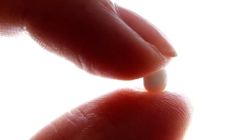 Primer plano de una pequeña píldora blanca sostenida entre la yema del pulgar y el índice, contra un fondo blanco brillante y desenfocado