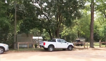 Vista exterior de una zona rural con un camión pickup blanco y un SUV plateado estacionados en un camino de tierra, rodeados de árboles y edificios