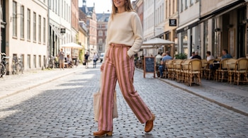 Mujer sonriendo camina por una calle empedrada, lleva suéter crema, pantalón ancho de rayas rosas y café claro, zuecos y bolso de tela.