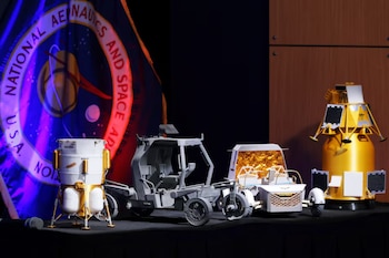 Tres modelos de naves espaciales y vehículos lunares se exhiben en una mesa oscura frente a una bandera de la NASA con iluminación azul