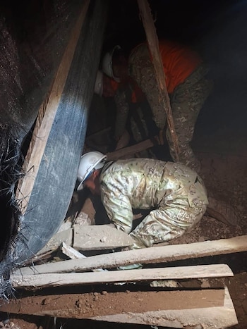 Dos personas vestidas con uniforme de camuflaje y casco blanco están entre escombros, tablas de madera y tierra en un espacio oscuro
