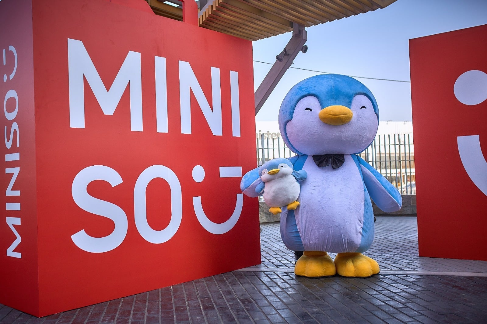 La mascota de Miniso, un pingüino azul y blanco, posa con un peluche de pingüino más pequeño frente a un gran letrero rojo de la marca en Chile