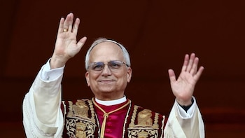 Primer plano del Papa Leo XIV, un hombre de mediana edad con gafas, sonriendo y agitando ambas manos. Viste una sotana blanca y una vestimenta roja y dorada