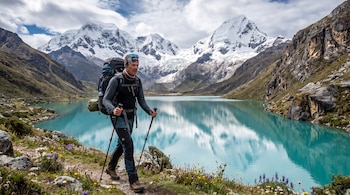 Hombre con mochila de excursionismo y bastones camina por un sendero de tierra junto a una laguna turquesa con montañas nevadas y un cielo nublado.