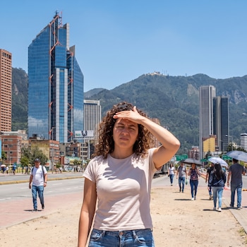 Una mujer con la mano en la frente camina por una calle con peatones, edificios altos y montañas de fondo bajo un cielo despejado.