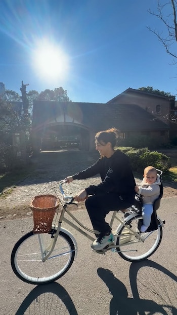 Una mujer conduce una bicicleta beige con canasto de mimbre con un bebé en un asiento posterior en una calle soleada