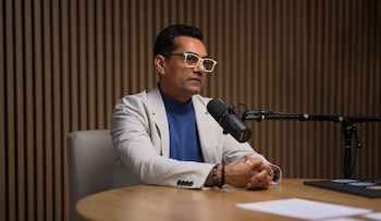 Hombre de mediana edad con gafas y blazer claro sentado frente a un micrófono en una mesa de madera, con las manos entrelazadas y un fondo de pared de madera texturizada