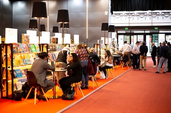 Vista interior de un espacio amplio con alfombra roja, personas conversando, sentadas y de pie junto a estanterías de libros iluminadas