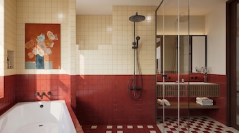 Baño con azulejos rojos y crema en las paredes, bañera blanca, ducha negra, cuadro decorativo y mampara de vidrio.