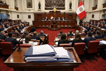 Salón principal del Congreso de Perú con bancas rojas, mesa directiva y parlamentarios sentados. Una bandera peruana, un micrófono y documentos en un atril.