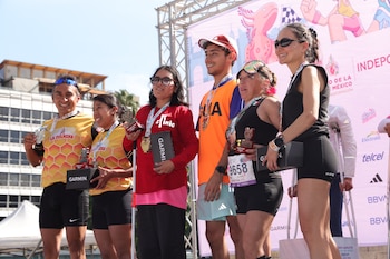 Seis personas en un podio con medallas, trofeos y cajas negras con el logo Garmin. Hay una persona con bastón blanco. Fondo con pancarta rosa