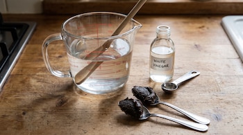 Jarra medidora con agua y cuchara de madera, botella de vinagre blanco, dos cucharas con jabón oscuro y cuchara medidora de metal sobre madera.