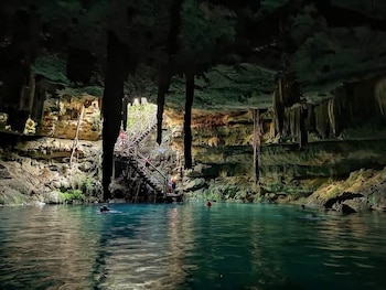 Cueva con agua turquesa, estalactitas colgantes, paredes rocosas, escalera de madera descendente y varias personas nadando