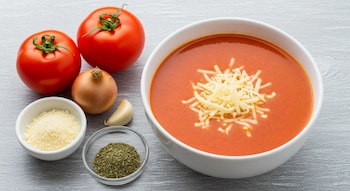 Un tazón blanco con sopa de tomate y queso rallado, dos tomates rojos, una cebolla, un diente de ajo, un cuenco con queso rallado y otro con orégano seco.