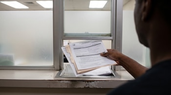 Mano de una persona entregando una pila de documentos en una ventanilla de servicio, con una superficie de mostrador y una ventana esmerilada de fondo.