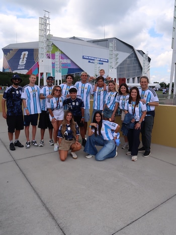 Grupo de personas con camisetas de la selección argentina frente a un estadio con el logo FIFA World Cup 2026. Un bebé en hombros de un adulto