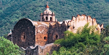 Antiguo edificio de piedra con cúpula y cruz, muros desgastados, rodeado de árboles y vegetación densa, con una colina boscosa
