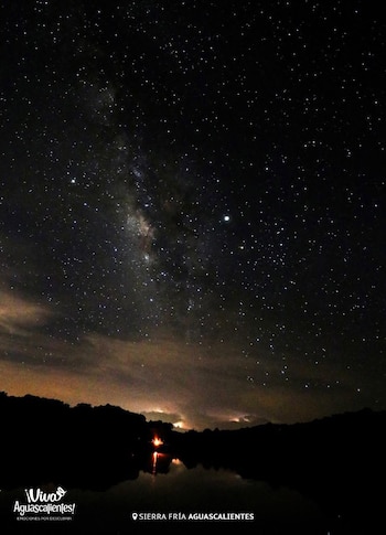 Cielo nocturno oscuro con innumerables estrellas y la Vía Láctea. Nubes bajas. Silueta de montañas y árboles. Fuego en la orilla de un lago, con su reflejo en el agua