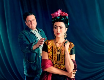 Mujer con flores rosas en el cabello, blusa bordada y joyas, junto a hombre con chaqueta de mezclilla. Fondo de tela azul oscuro