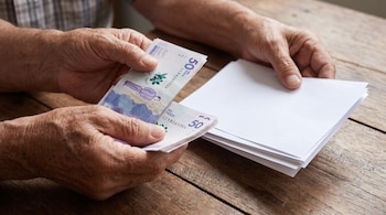 Primer plano de manos envejecidas sobre una mesa de madera. Una mano sostiene billetes colombianos de 50 mil pesos, y la otra, una pila de papeles en blanco.