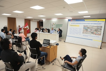 Vista general de un salón con personas sentadas en sillas, un orador en un podio con bandera peruana y un panel de expertos, frente a una pantalla de proyección